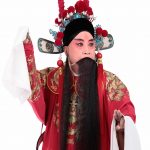 在那纯白的背景前，身穿红色戏服的他，头戴华丽帽饰，仿佛从历史的长河中走来，带着岁月的痕迹和故事。他站在那里，就像一座山，稳重而不可动摇。他的每一个动作，每一个表情，都充满了力量，让人感受到京剧艺术的魅力。他的眼神中透露出一种深邃与思考，思索着人生的大道理。